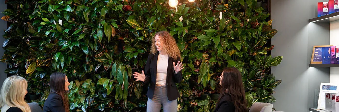 A woman presents in front of a green plant wall, engaging in a discussion about digital transformation and system integration, representing Akkodis' Digital Backbone Services. A woman presents in front of a green plant wall, engaging in a discussion about digital transformation and system integration, representing Akkodis' Digital Backbone Services.