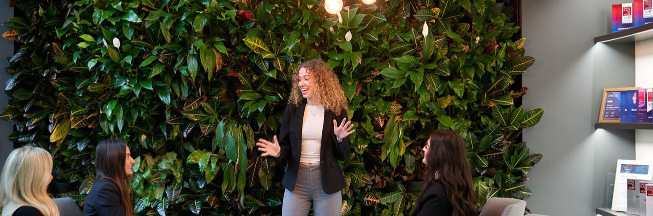 A woman presents in front of a green plant wall, engaging in a discussion about digital transformation and system integration, representing Akkodis' Digital Backbone Services. A woman presents in front of a green plant wall, engaging in a discussion about digital transformation and system integration, representing Akkodis' Digital Backbone Services.