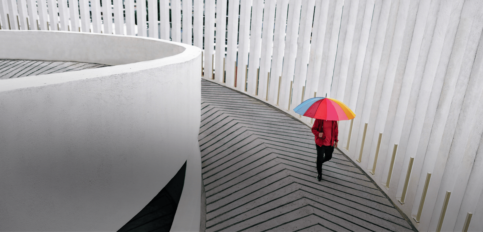 Una persona con un ombrello colorato cammina su un percorso curvo tra pareti di architettura moderna bianca.